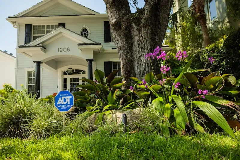 An ADT security yard sign stands in a lush green lawn in front of a white two-story house with a large tree and purple flowers.